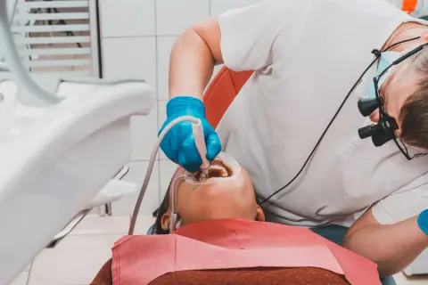 Dentysta w niebieskich rękawiczkach wykonuje piaskowanie zębów pacjentowi, używając specjalistycznego sprzętu.