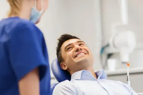 Uśmiechnięty mężczyzna w gabinecie dentystycznym, rozmawiający z higienistką. Pytanie o fluoryzacja zębów skutki uboczne.
