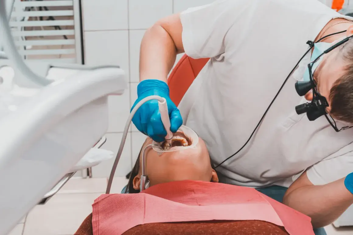 Dentysta w niebieskich rękawiczkach wykonuje piaskowanie zębów pacjentowi, używając specjalistycznego sprzętu.